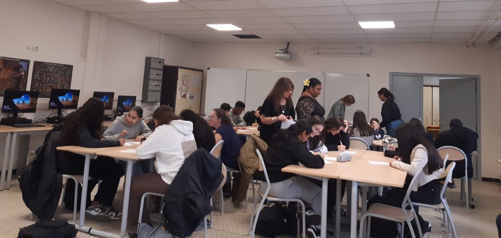 Plusieurs ilots avec des élèves qui travaillent en groupe dans une classe, avec les professeurs debouts