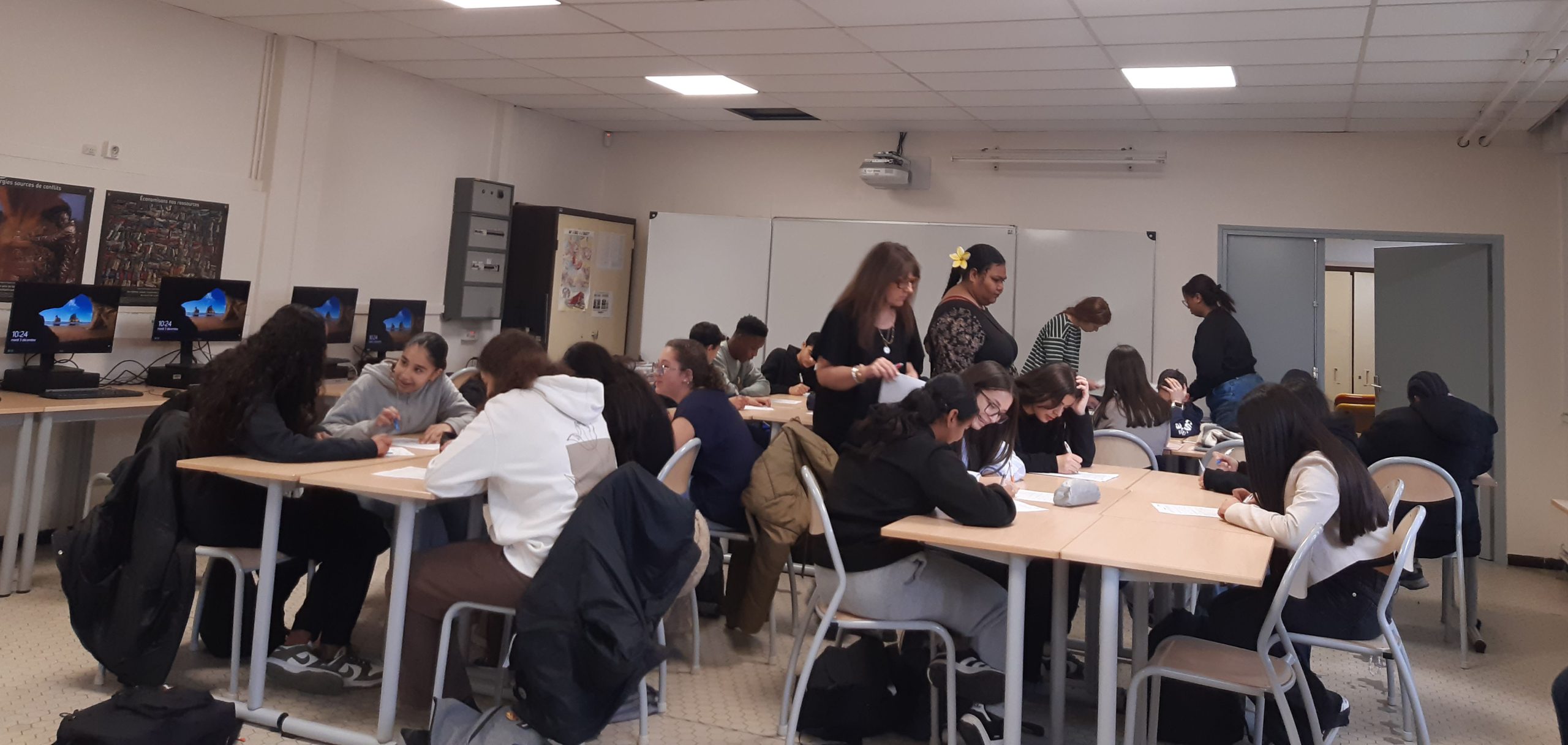 Plusieurs ilots avec des élèves qui travaillent en groupe dans une classe, avec les professeurs debouts